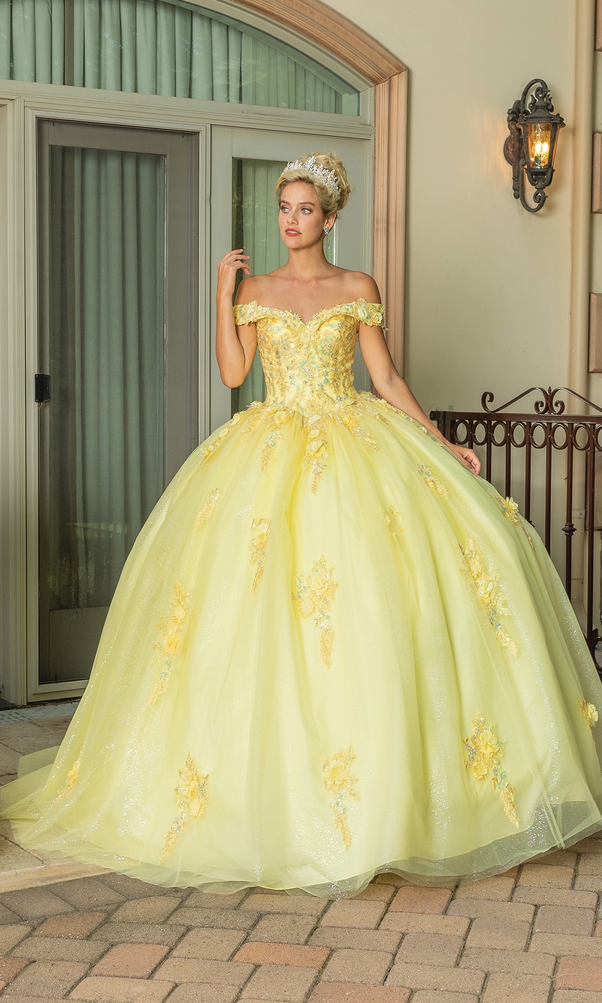 A woman in the Quinceanera Dress 1760 By Dancing Queen, a yellow off-the-shoulder gown with floral details and a sweetheart neckline, wears a tiara and stands before elegant glass doors and a wrought iron railing.